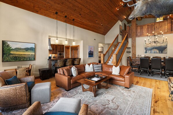 A spacious living room with a large brown leather sectional sofa, wooden coffee table, and a patterned rug. A staircase with wooden railings leads to an upper level.