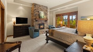 A bedroom with a stone fireplace, a bed, a TV, and a window with a view of trees. - Ski-In/Ski-Out Luxury | Cowboy Heaven Retreat (Big Sky)