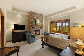 A bedroom with a stone fireplace, a bed, a TV, and a window with a view of trees.