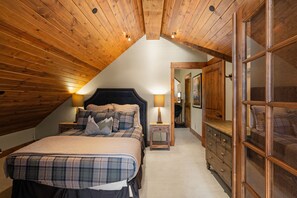 Um quarto de sótão aconchegante com teto inclinado, uma cama grande com roupa de cama xadrez, uma mesa de cabeceira com abajur e um aparador de madeira.