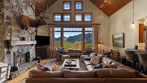 A spacious living room with a stone fireplace, leather sofa, and a large window offering a mountain view. - Ski-In/Ski-Out Luxury | Cowboy Heaven Retreat (Big Sky)