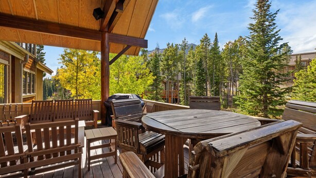 An outdoor wooden deck with a round table and benches, surrounded by trees and a mountain view. - Ski-In/Ski-Out Luxury | Cowboy Heaven Retreat (Big Sky)
