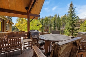 An outdoor wooden deck with a round table and benches, surrounded by trees and a mountain view.