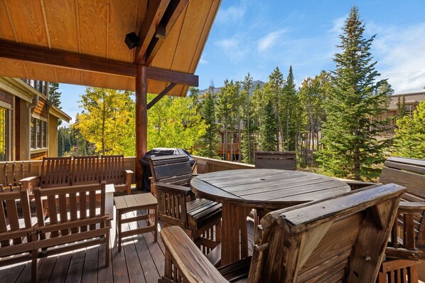 An outdoor wooden deck with a round table and benches, surrounded by trees and a mountain view.