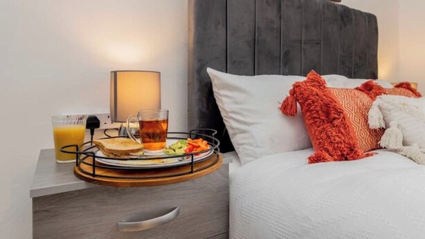 Una cama bien hecha con ropa blanca y una almohada roja, una mesita de noche con una bandeja de desayuno que incluye tostadas, té y fruta, y una lámpara que proporciona una iluminación suave.