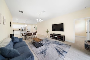 Una sala de estar moderna con un sofá, una mesa de centro y un mueble para televisión. Se observa un comedor al fondo con una mesa y sillas.