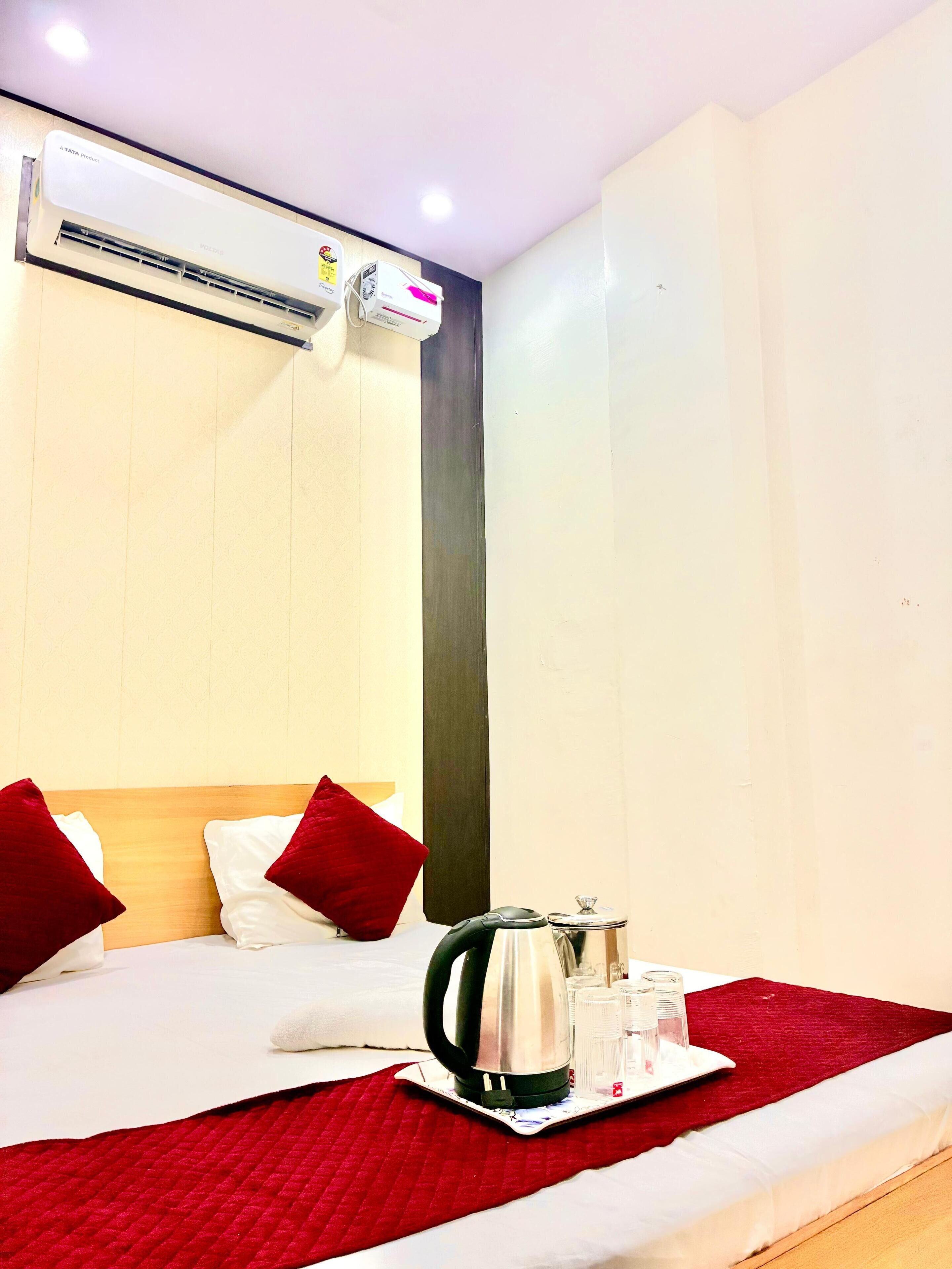 A hotel room with a bed, red and white pillows, a tray with a kettle, glasses, and a cup. There is an air conditioning unit on the wall.
