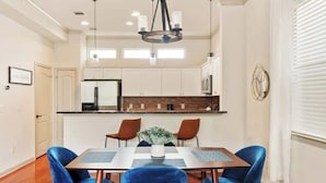 Uma sala de jantar com uma mesa retangular, quatro cadeiras azuis, um lustre e uma cozinha ao fundo.