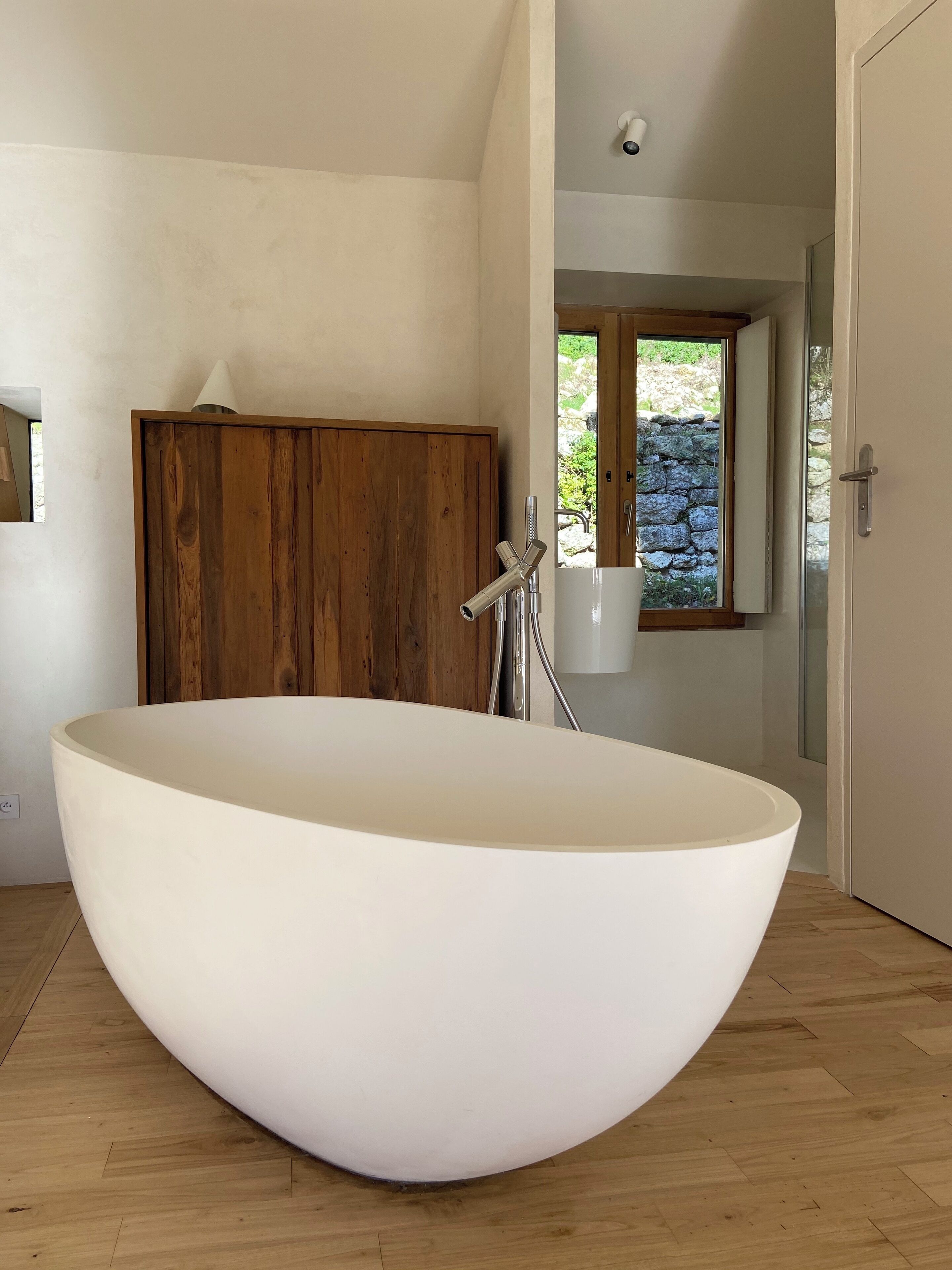 A modern bathroom with a large freestanding bathtub, wooden paneling, and a stone wall visible through a window.