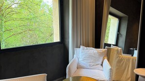 Una habitación de hotel moderna con un ventanal grande, un sofá blanco, una mesita redonda y un espejo.