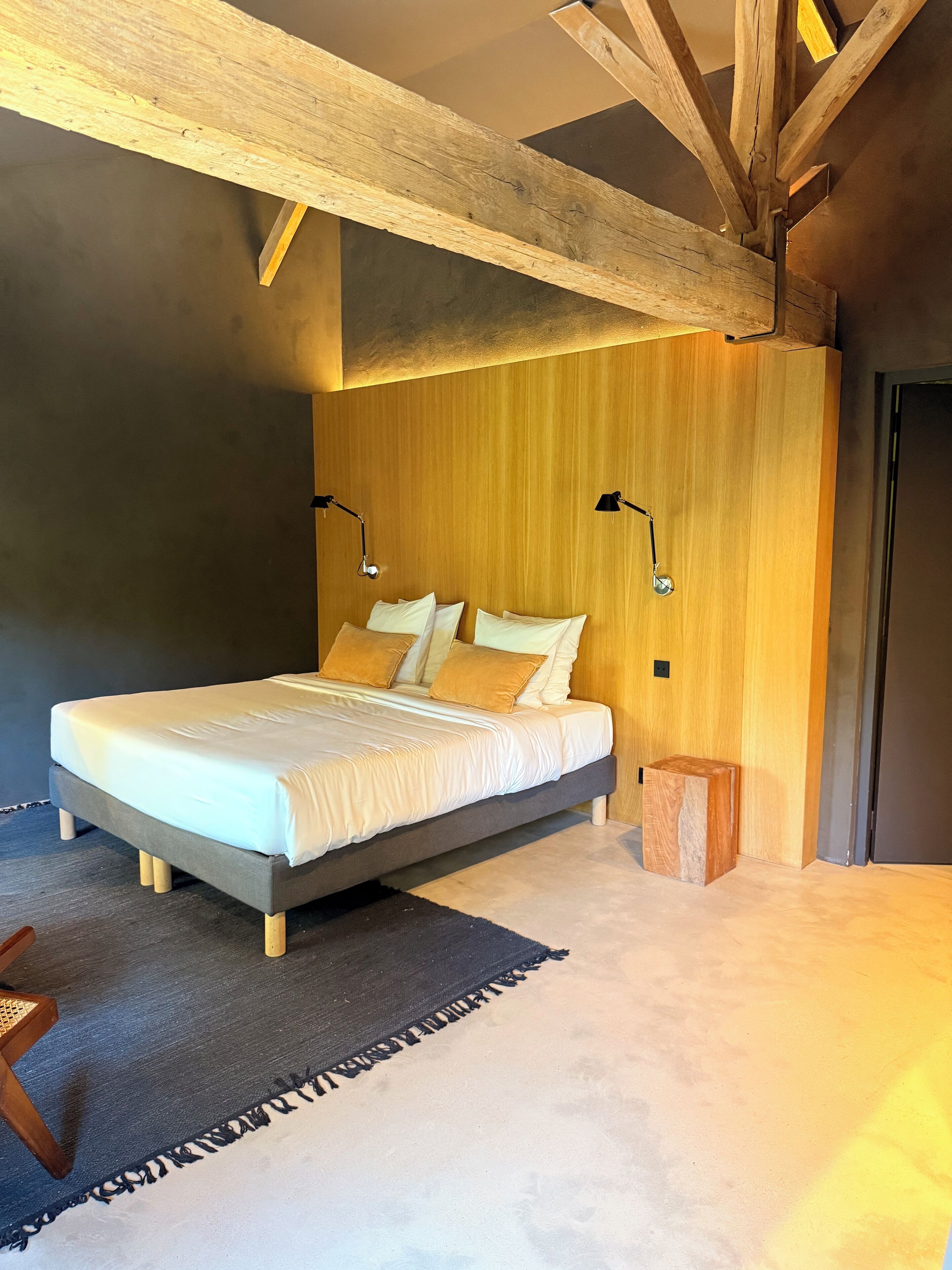 Een slaapkamer met een bed, een houten hoofdbord, twee bedlampen, een klein houten krukje en een donker vloerkleed.