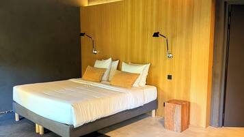 Een slaapkamer met een bed, een houten hoofdbord, twee bedlampen, een klein houten krukje en een donker vloerkleed.