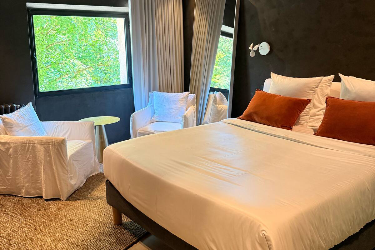 A modern bedroom with a large bed, a small table, two chairs, a mirror, and a window with a view of greenery.