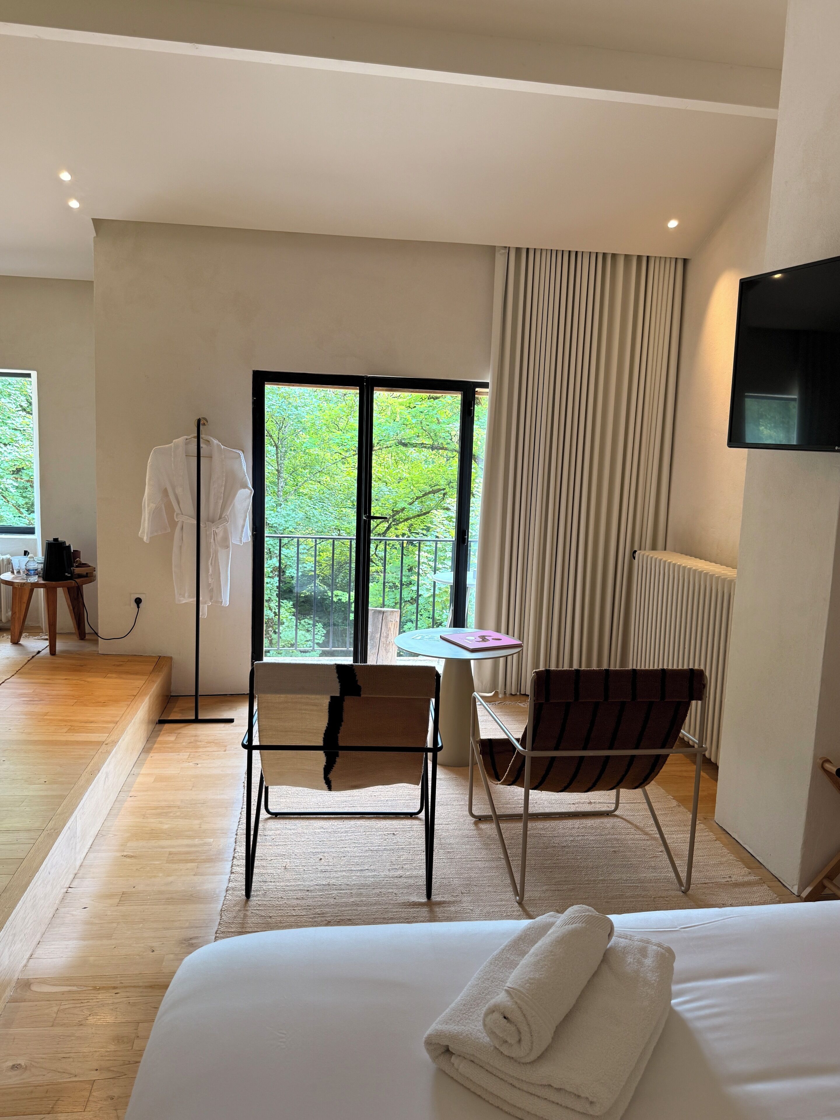 Habitación de hotel con cama, dos sillas, una mesa, perchero para bata y televisor.