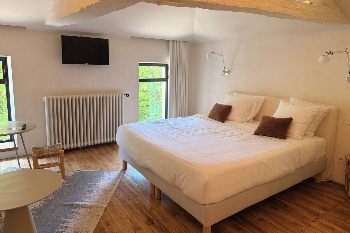 A bedroom with a large bed, wooden floor, a radiator, a TV on the wall, and a window with curtains.