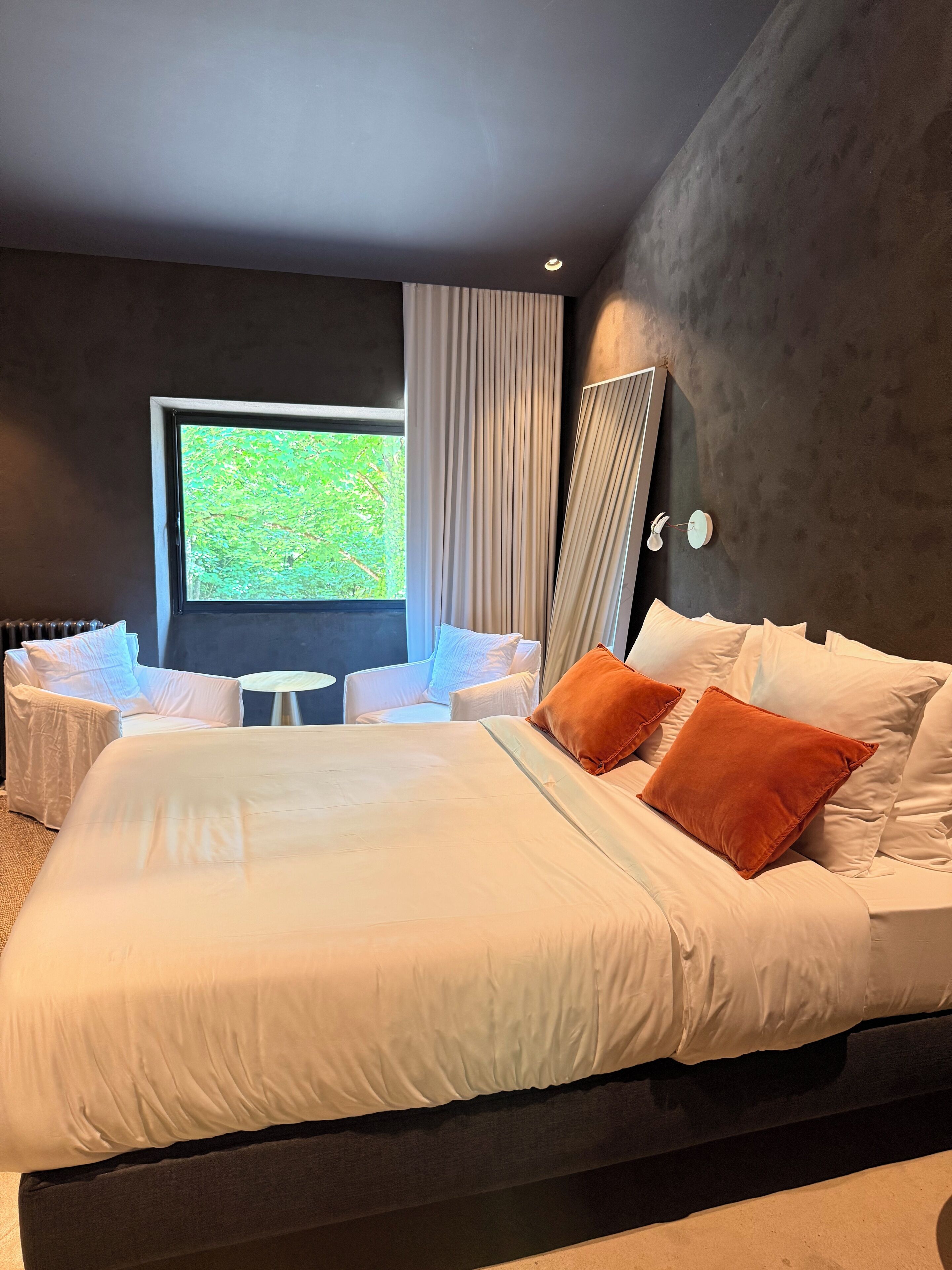 A modern bedroom with a large bed, white bedding, and a view of greenery through a window.