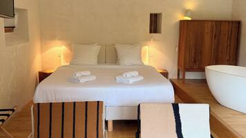 Habitación de hotel con una cama grande, dos mesitas de noche, un armario de madera, una bañera blanca y dos sillones a rayas.