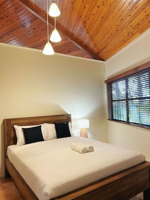 A bedroom with a wooden ceiling, a bed with white bedding, two bedside lamps, and a window with blinds.