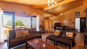 Une pièce confortable, aménagée dans le style d’un chalet, avec un canapé, deux fauteuils, une table basse et une table à manger. Une grande fenêtre offre une vue sur l’extérieur.