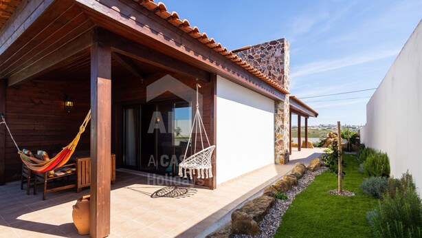 Une maison en bois avec un patio carrelé, un hamac et un jardin agrémenté de rochers et de plantes.