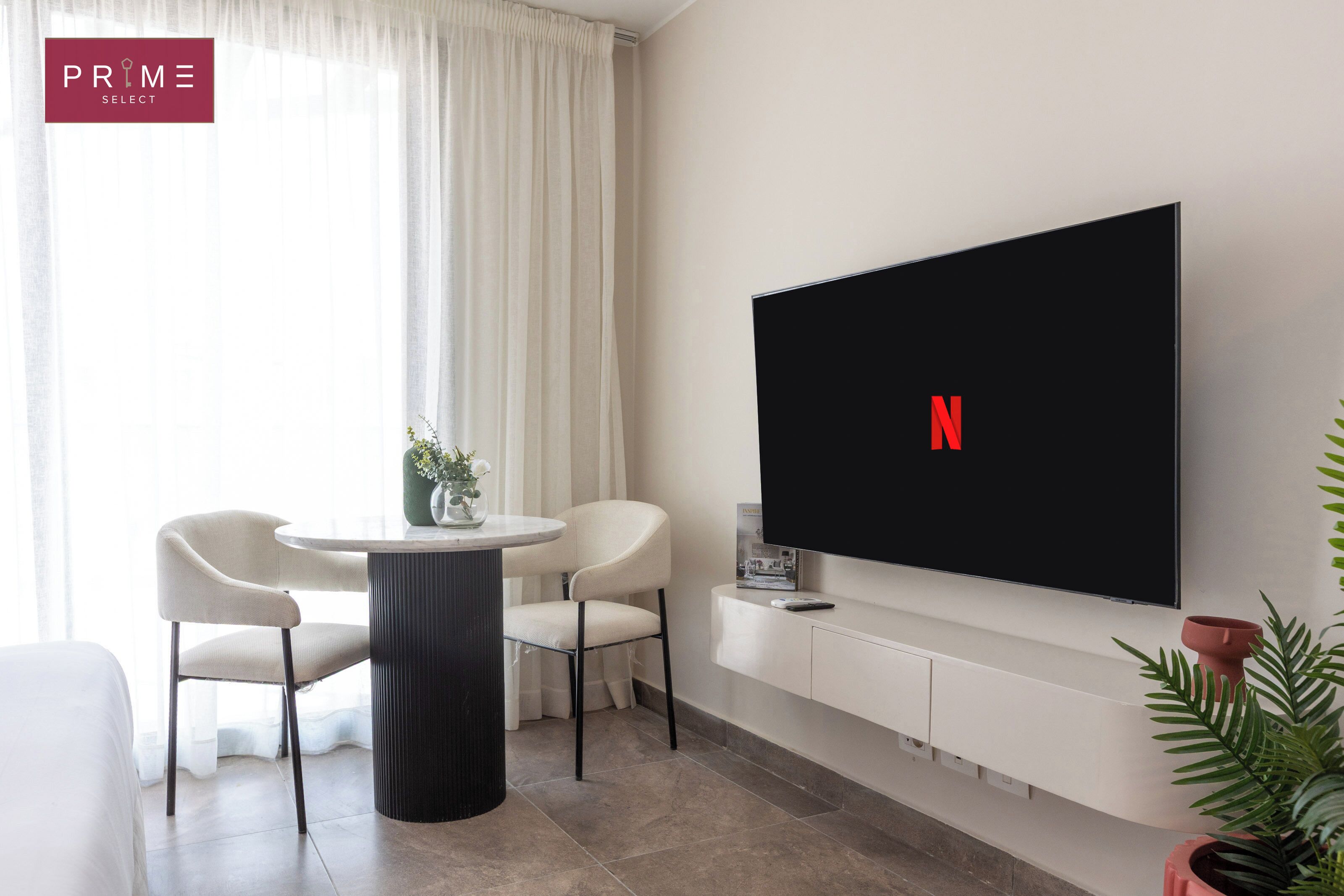 A modern living room with a flat-screen TV mounted on the wall, a round dining table with two chairs, a potted plant, and a large window with sheer curtains.