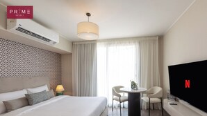 A hotel room with a bed, a TV, a desk with chairs, and a large window with curtains.