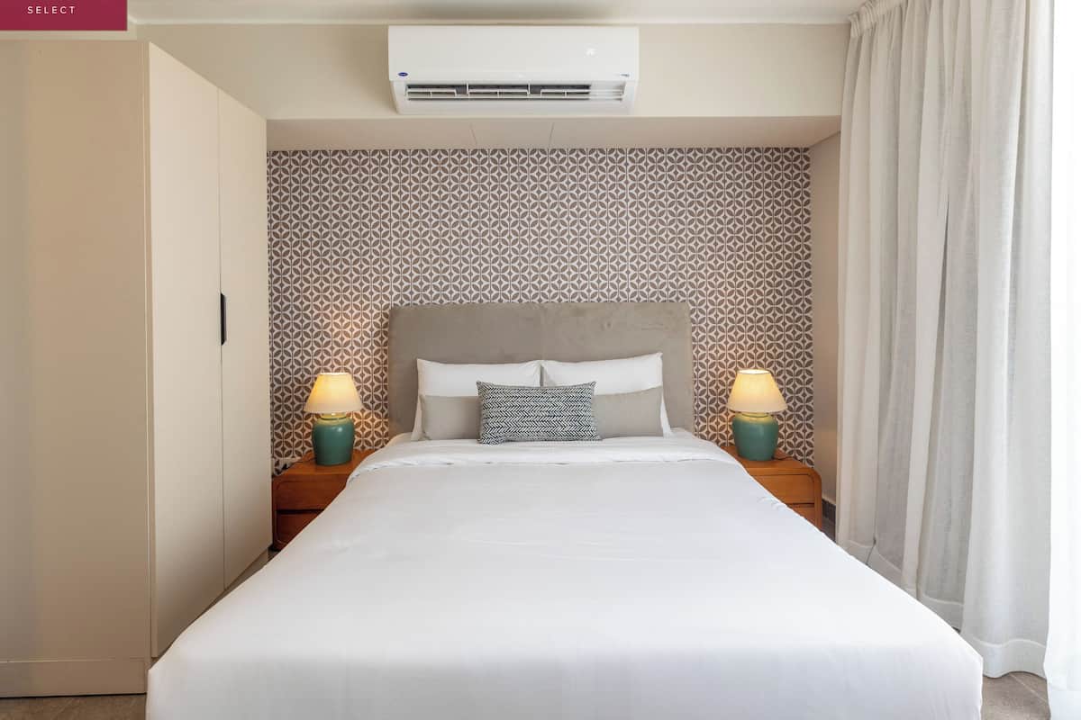 Une chambre d’hôtel avec un grand lit, deux tables de chevet avec des lampes, un papier peint à motifs et un appareil de climatisation placé au-dessus du lit.