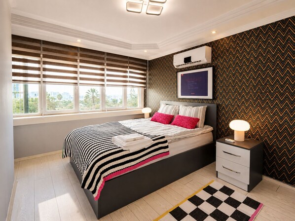 Une chambre moderne avec un grand lit, des tables de chevet, une fenêtre avec des stores, un climatiseur fixé au mur et un mur décoré d’un motif.