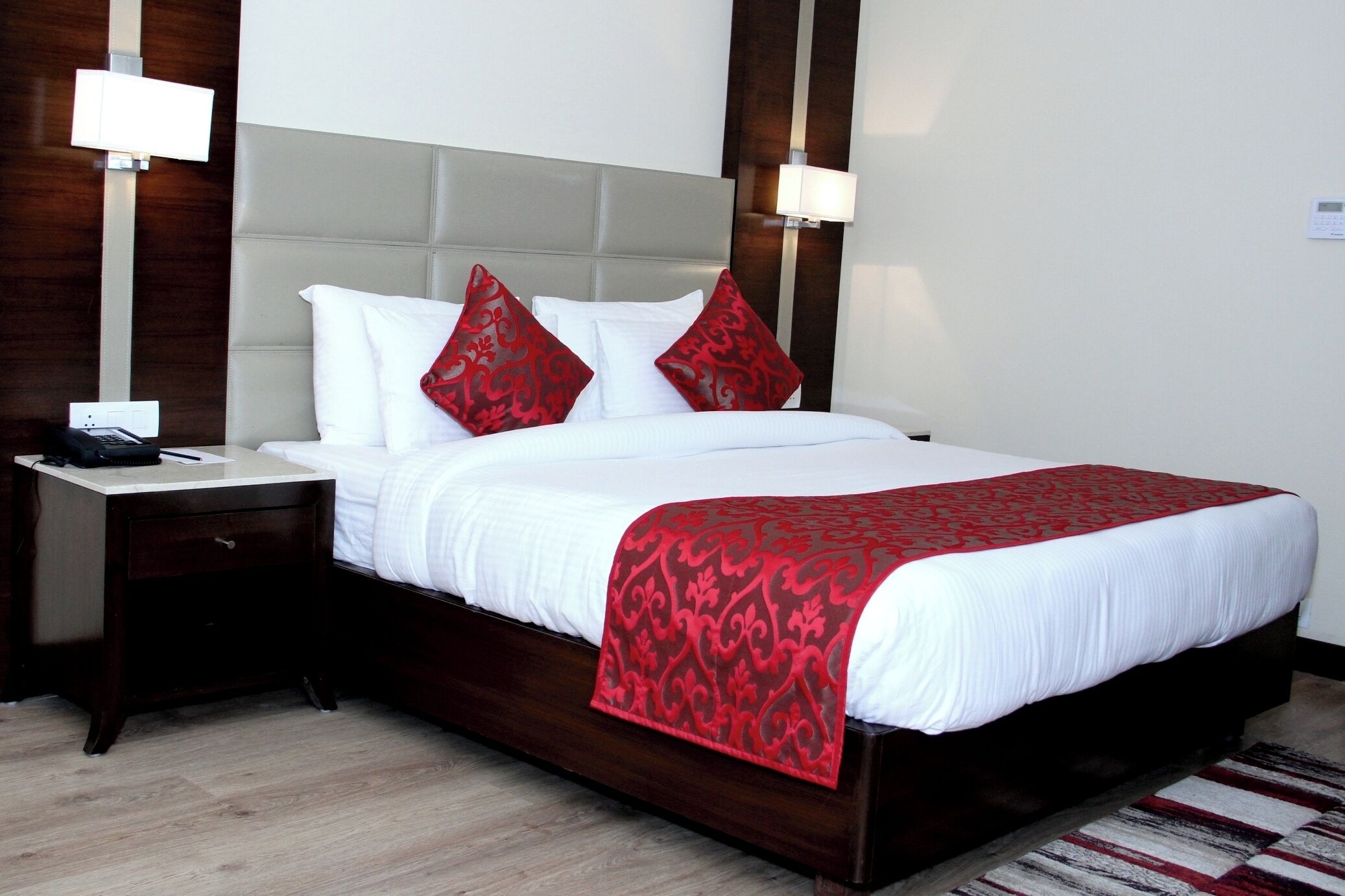 Una cama bien tendida con ropa blanca y roja, una mesita de noche de madera con una lámpara y un cabecero moderno.