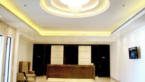 A reception area with a modern design, featuring a central chandelier, a reception desk, and a seating area with chairs.