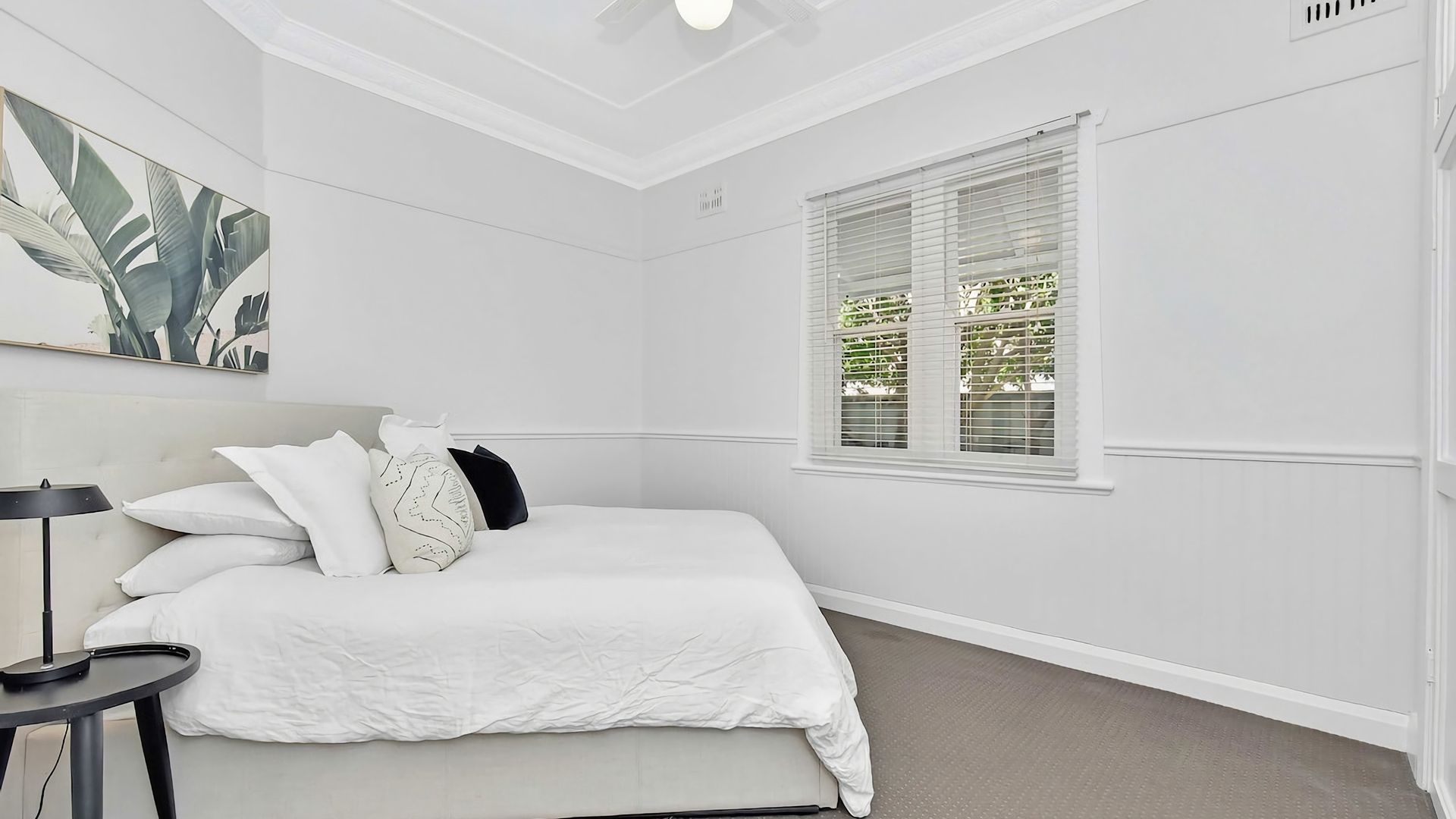 A bedroom with a bed, pillows, a bedside table, a lamp, and a window with blinds.