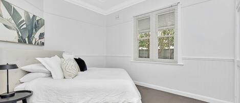 A bedroom with a bed, pillows, a bedside table, a lamp, and a window with blinds.