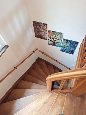 Un escalier en bois, doté d’une rampe, mène à un palier où sont accrochées deux œuvres d’art représentant des arbres.