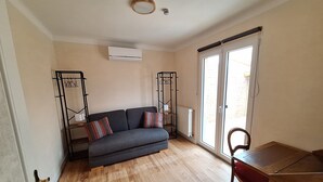 Una habitación con un sofá gris, una mesa de comedor de madera y una silla. Hay un radiador en la pared y una ventana con vistas al exterior.