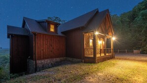 Una cabaña de madera con cimientos de piedra, un porche iluminado y un patio cercado durante la noche.