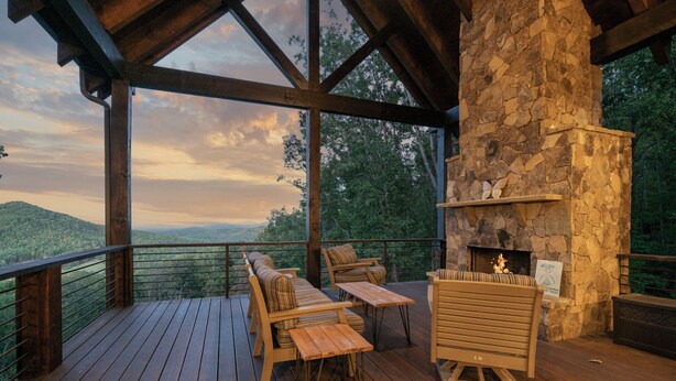 Una terraza de madera con mobiliario exterior, una chimenea de piedra y una vista panorámica de las montañas.