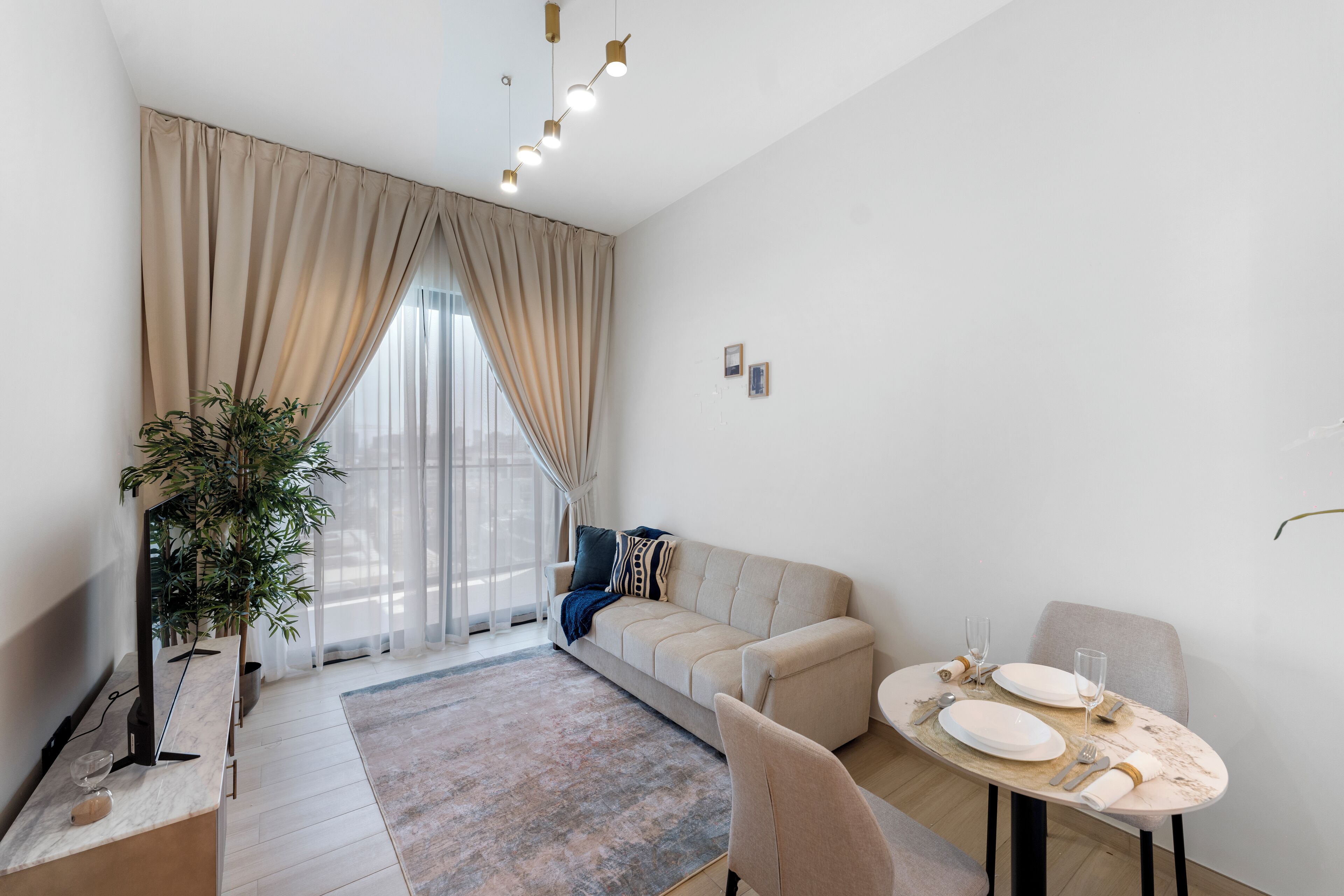Een moderne woonkamer met een bank, een eettafel gedekt voor twee personen, een groot raam met gordijnen en een potplant.