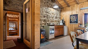 Interior acogedor de una cabaña con pared de piedra, vigas de madera, comedor y cocina con estufa.