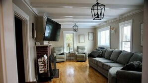 A living room with a grey sofa, two armchairs, a TV mounted on the wall, and a fireplace. - Welcoming 5-bedroom farmhouse in Rangeley (Rangeley)