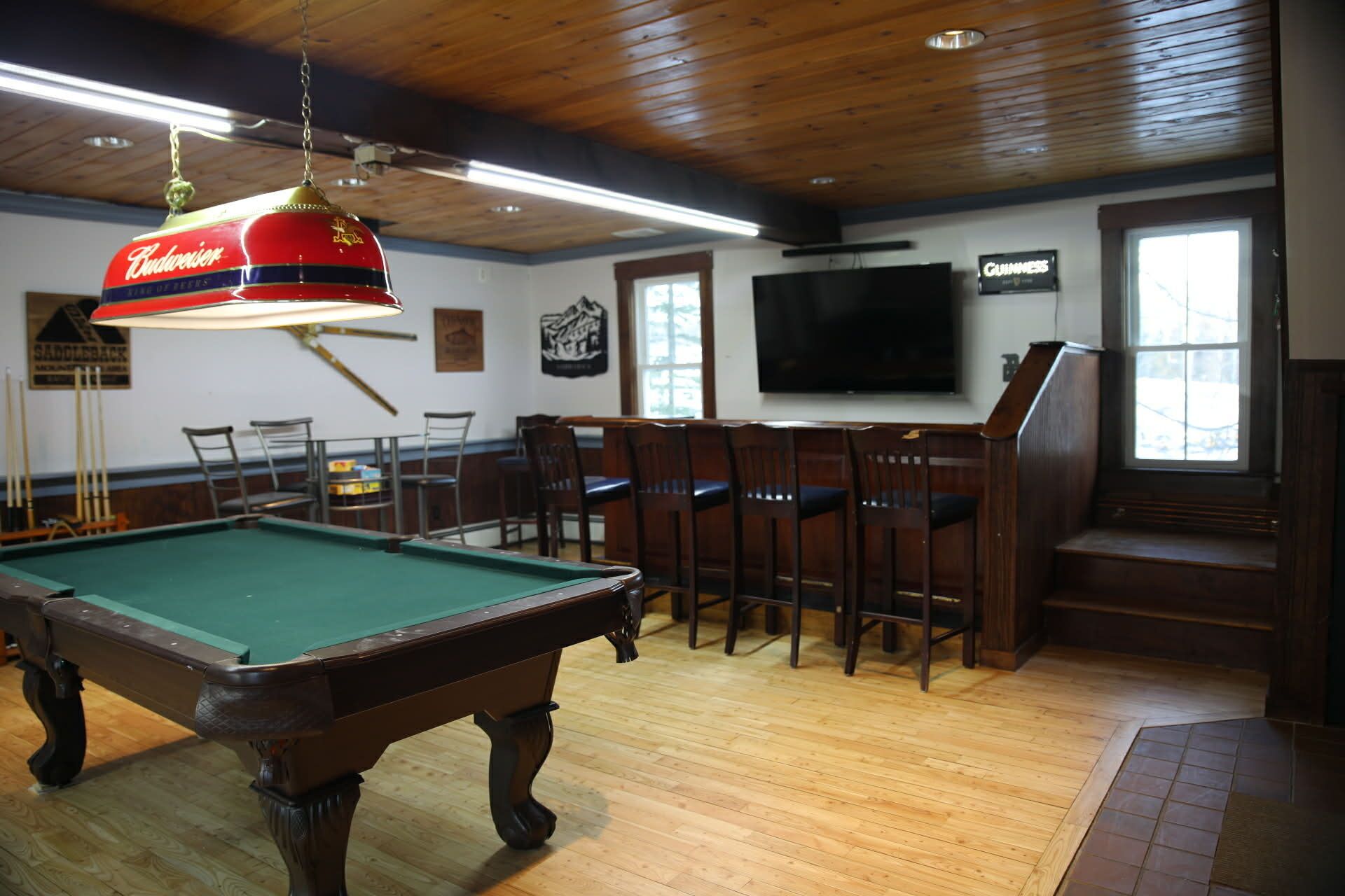 Een pooltafel met een hanglamp, houten vloer, een aan de muur bevestigde televisie en een bar met stoelen.