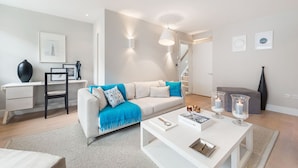 A modern living room with a sofa, coffee table, and a desk with a chair. There are wall-mounted lights and a framed picture on the wall. - Prime Knightsbridge Mews-4 bed House (London)