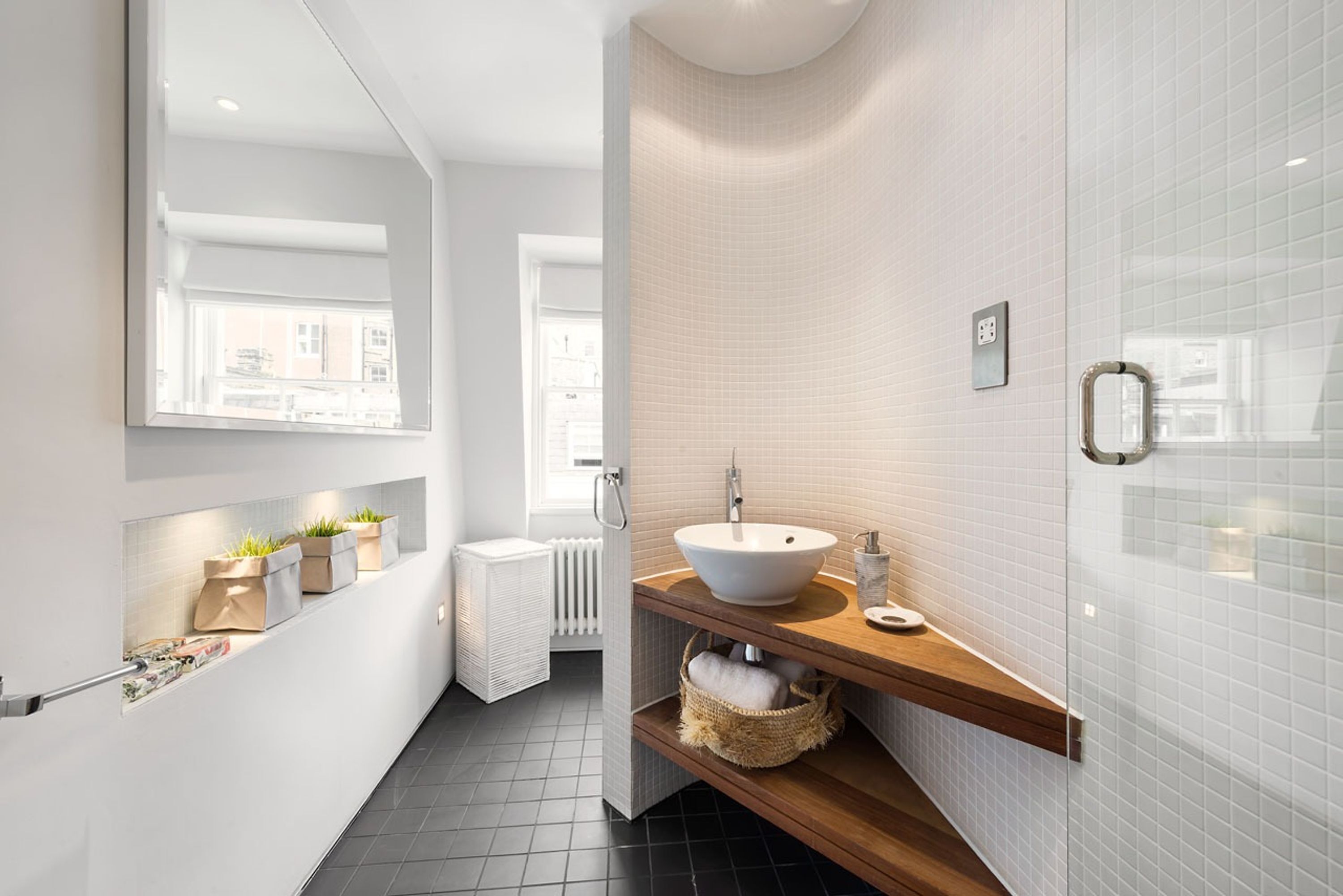 Une salle de bain moderne avec un grand miroir, des murs recouverts de carreaux blancs, un meuble-lavabo en bois et une fenêtre laissant entrer la lumière naturelle.