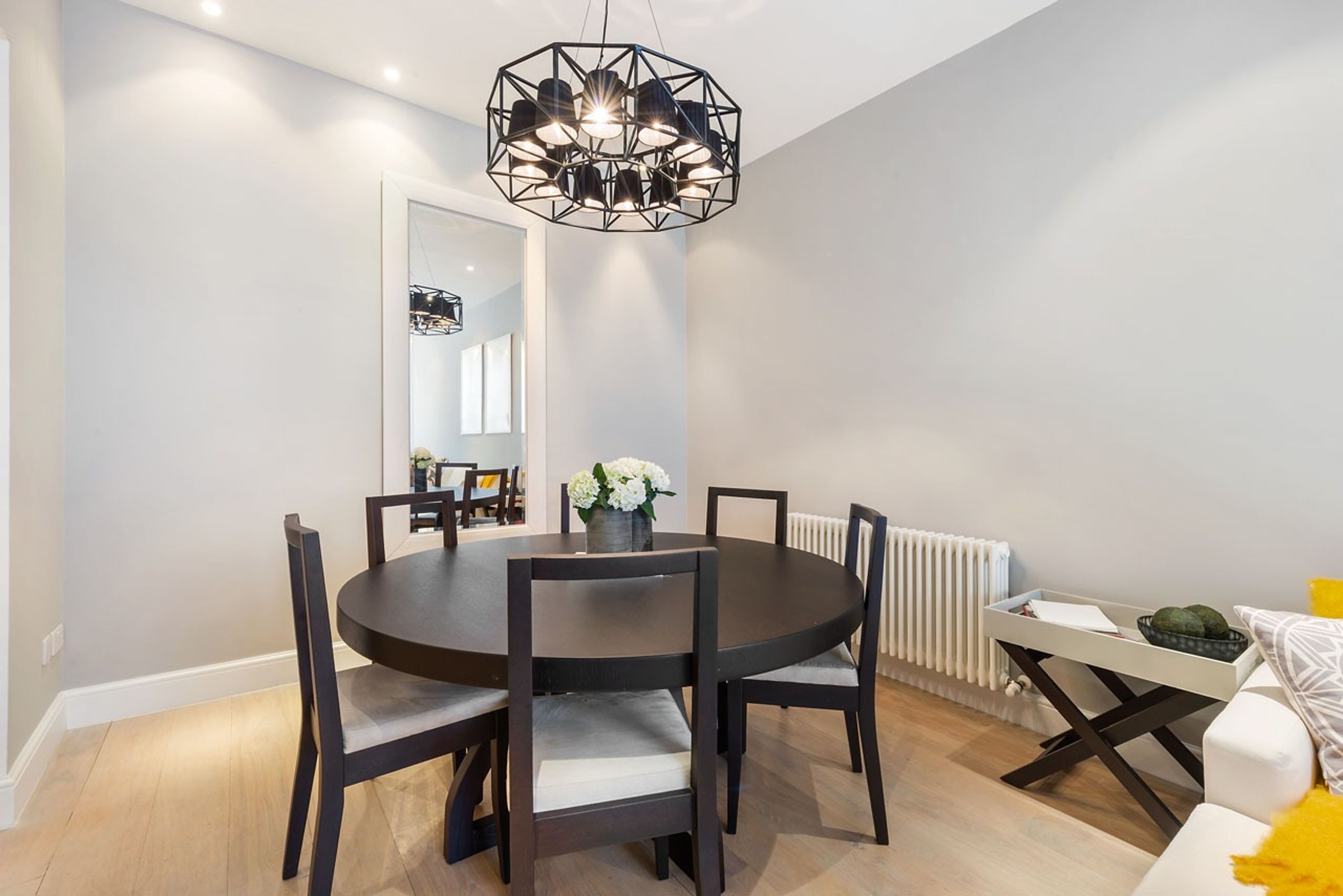A dining area with a round table, six chairs, a modern chandelier, and a white radiator.