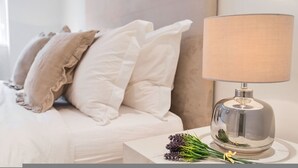 A neatly made bed with white and beige bedding, a clear glass lamp on a bedside table, and a bouquet of flowers. - Prime Knightsbridge Mews-4 bed House (London)