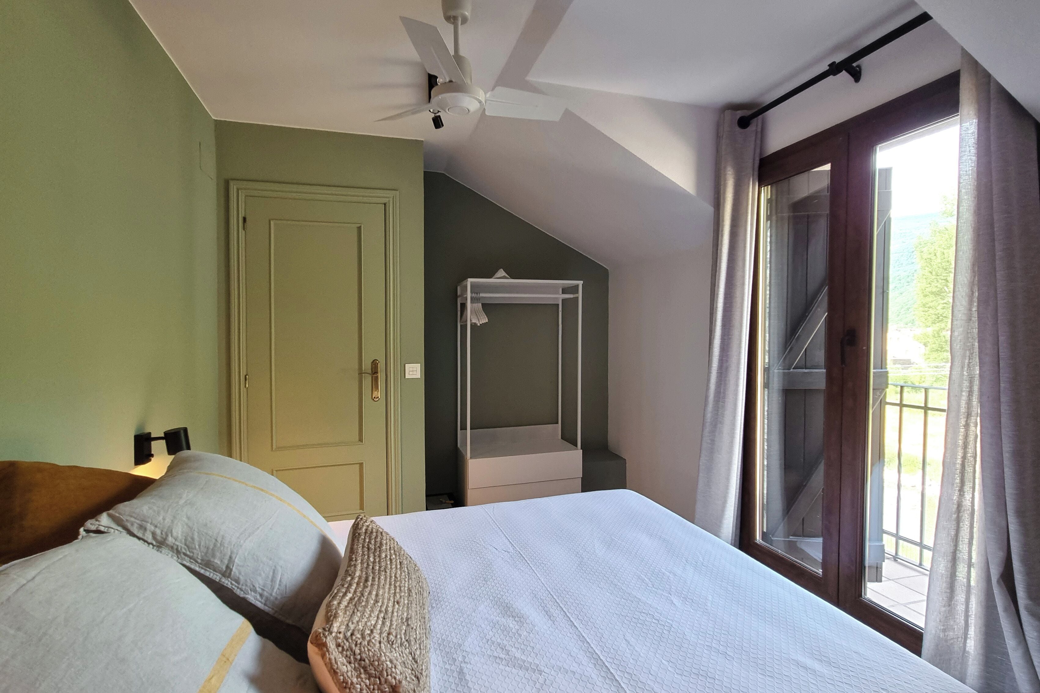 Une chambre à coucher comprenant un lit, un ventilateur de plafond, une fenêtre avec des rideaux, une armoire et un petit balcon.