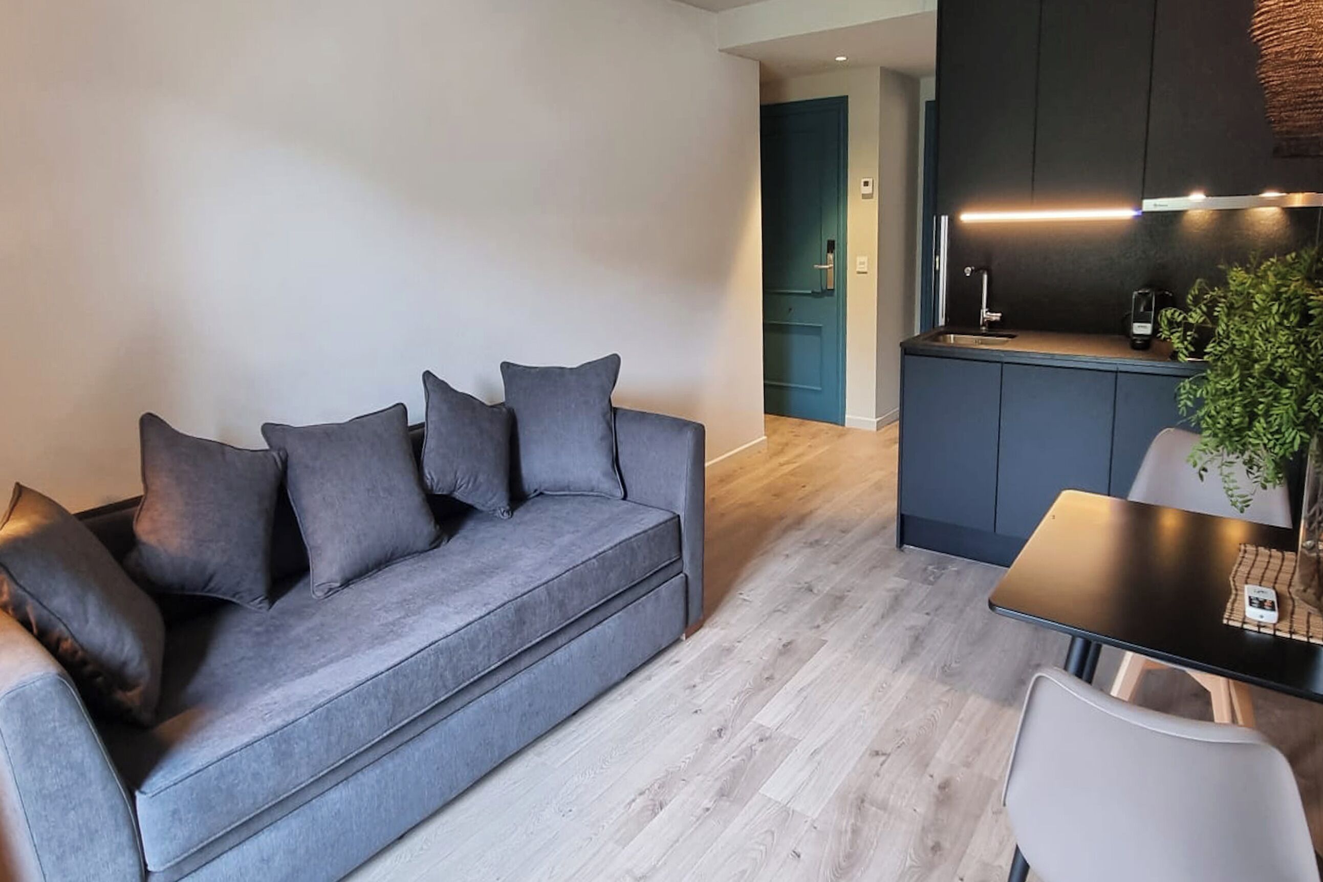 A modern living room with a grey sofa, a small dining table, and a kitchen area in the background.