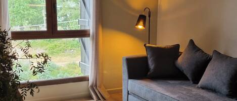 Una habitación con un sofá gris, una lámpara de pie, una ventana con cortinas y una planta.