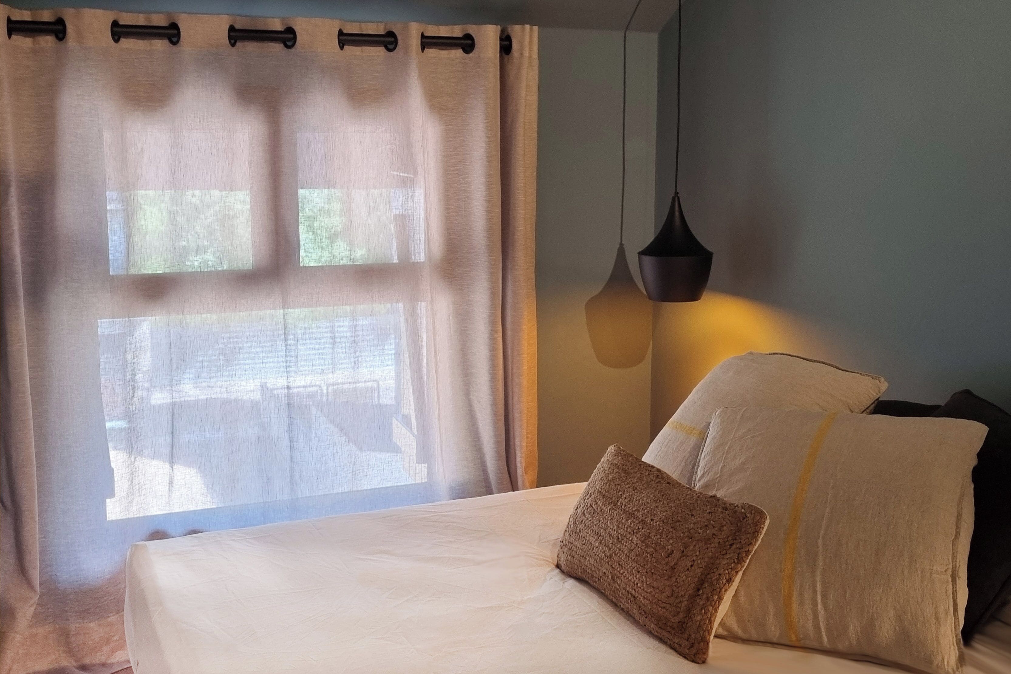 A neatly made bed with pillows and a hanging light fixture in a room with a window and curtains.