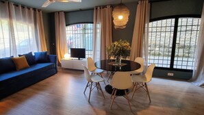 Una sala de estar moderna con un sofá azul, sillas blancas, una mesa redonda y ventanas grandes con cortinas.