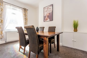 A dining area with a wooden table and four chairs, a radiator, a window with curtains, and a framed picture on the wall.